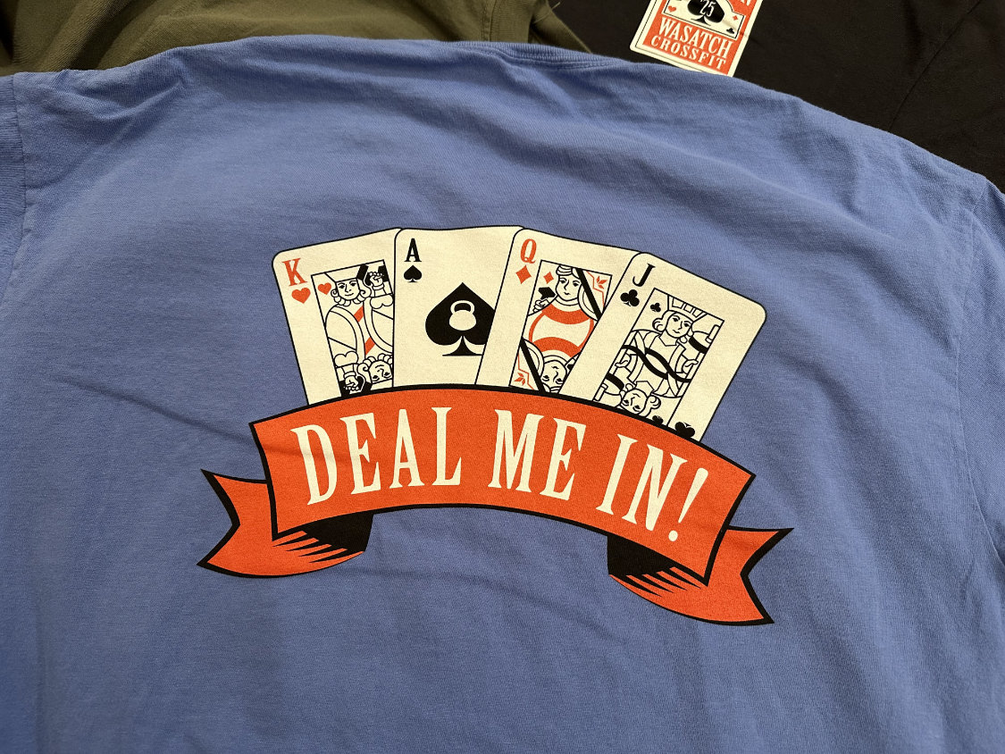 The back of a blue tee is decorated with a screen print featuring playing cards (King, Ace, Queen, and Jack), and a banner below stating, "DEAL ME IN!"